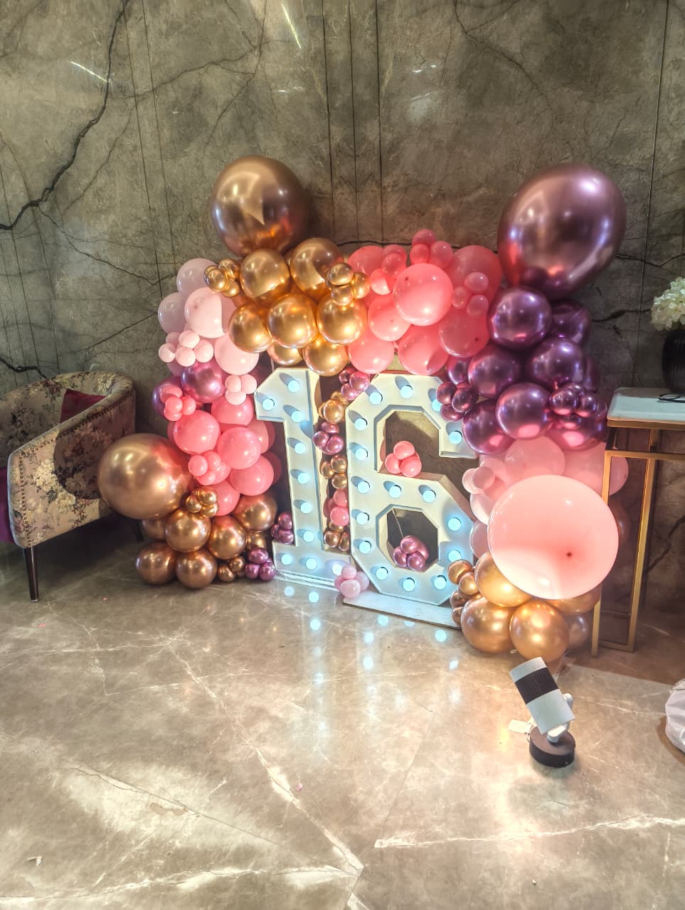A decorative party backdrop with a large illuminated marquee-style number 16 surrounded by clusters of pink, rose gold, and metallic gold balloons, set against a marble wall and floor.