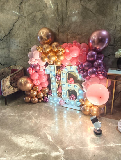 A decorative party backdrop with a large illuminated marquee-style number 16 surrounded by clusters of pink, rose gold, and metallic gold balloons, set against a marble wall and floor.